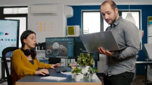 Project supervisor holding laptop and talking with engineer compering d industrial metallic pieces