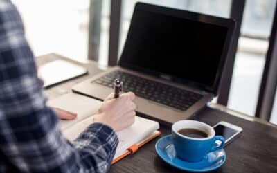 A man writing on a notebook in front of a laptop